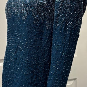 May include: A teal-colored, long-sleeved top with intricate beadwork. The top features a textured fabric and is adorned with small, shimmering beads that create a gradient effect. The sleeves and neckline are also embellished.