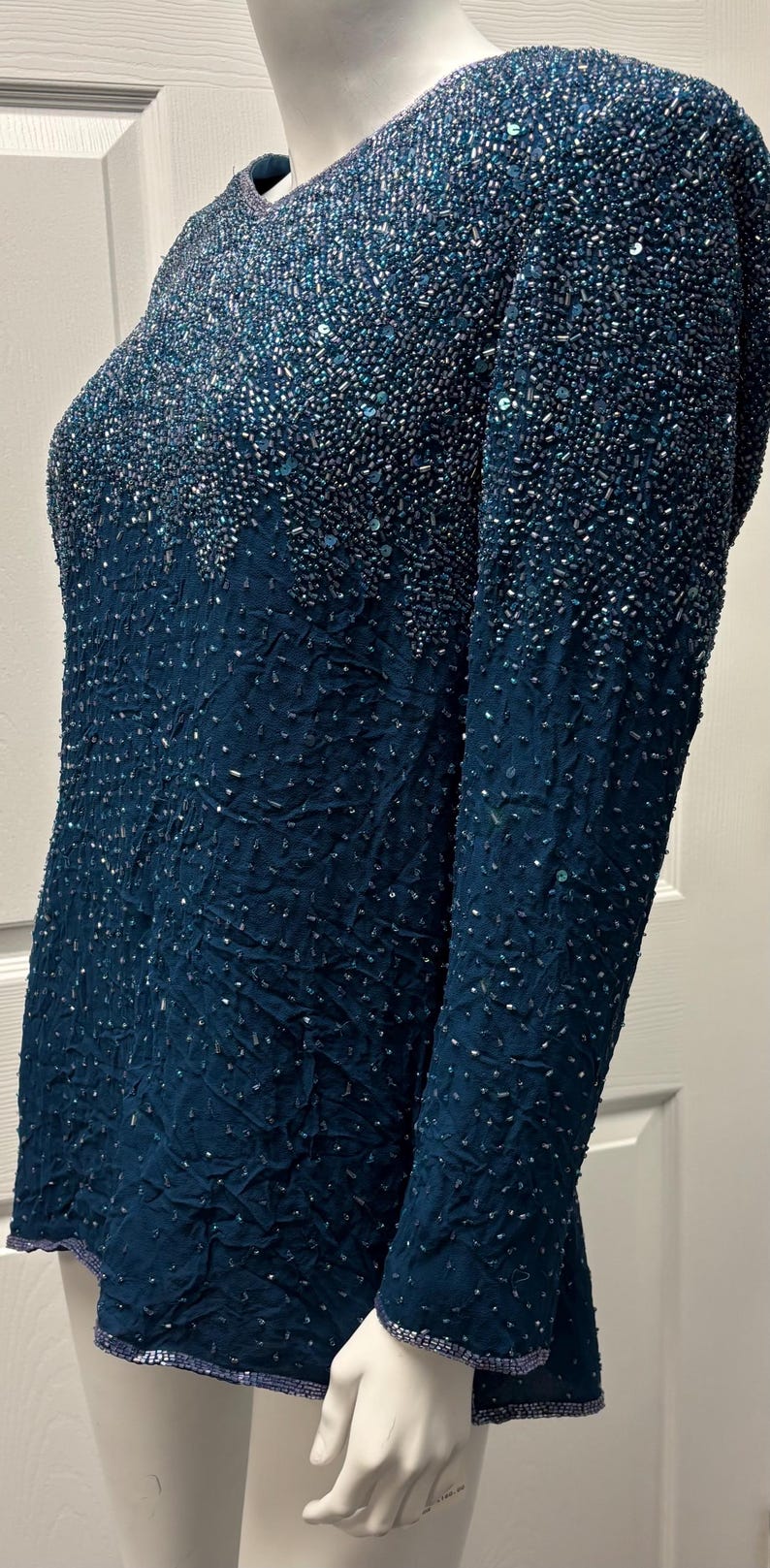 May include: A teal-colored, long-sleeved top with intricate beadwork. The top features a textured fabric and is adorned with small, shimmering beads that create a gradient effect. The sleeves and neckline are also embellished.