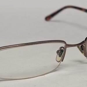 May include: A pair of eyeglasses with a brown metal frame and clear lenses. The temples are a darker brown with a tortoiseshell pattern near the hinges. The lenses are rectangular with a rounded bottom edge.