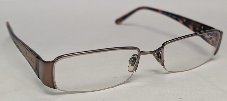 May include: A pair of eyeglasses with a brown metal frame and clear lenses. The temples are a darker brown with a tortoiseshell pattern near the hinges. The lenses are rectangular with a rounded bottom edge.