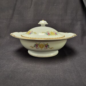 May include: A white porcelain serving bowl with a lid, featuring floral designs in yellow, pink, and blue. The bowl has a gold trim and a pedestal base. The lid is topped with a small gold finial.