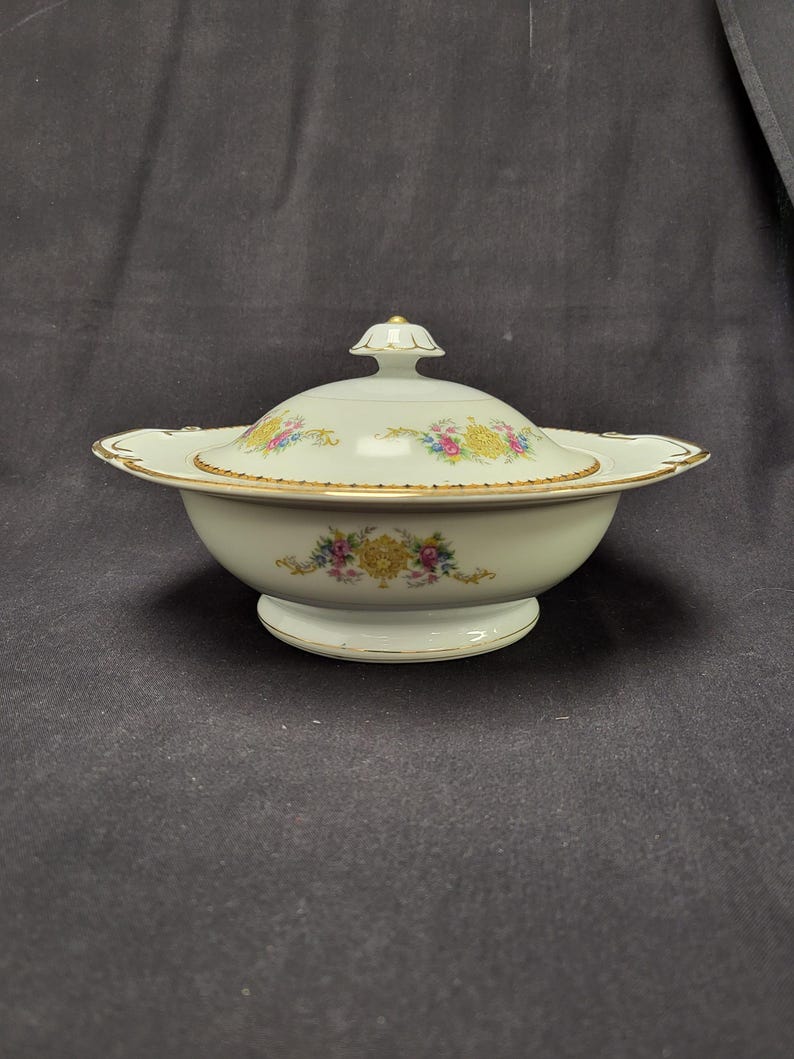 May include: A white porcelain serving bowl with a lid, featuring floral designs in yellow, pink, and blue. The bowl has a gold trim and a pedestal base. The lid is topped with a small gold finial.