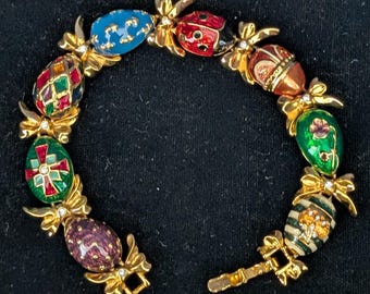 May include: A gold-toned bracelet featuring colorful, enameled egg-shaped charms. The charms are decorated with various patterns and designs, including a ladybug, crosses, and floral motifs. The bracelet is connected by gold-toned bows and small crystals.
