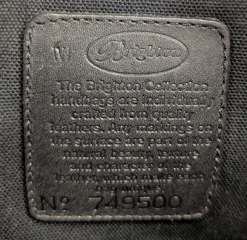 May include: A close-up of a black leather tag with the Brighton logo embossed at the top. The tag includes the text "The Brighton Collection handbags are individually crafted from quality leathers. Any markings on the surface are part of the natural beauty..." and the number 749500.