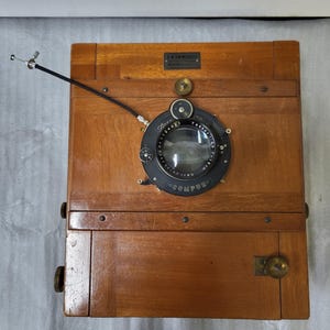 May include: A wooden large format camera with a black lens and a Compur shutter. The camera has a brass nameplate that reads "G.W. Emmerich Berlin-Steglitz".