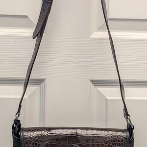 May include: A brown and black leather crossbody bag with a crocodile-textured flap. The bag has a silver buckle on the strap and a decorative silver and gold clasp. The bag is hanging from a white door.