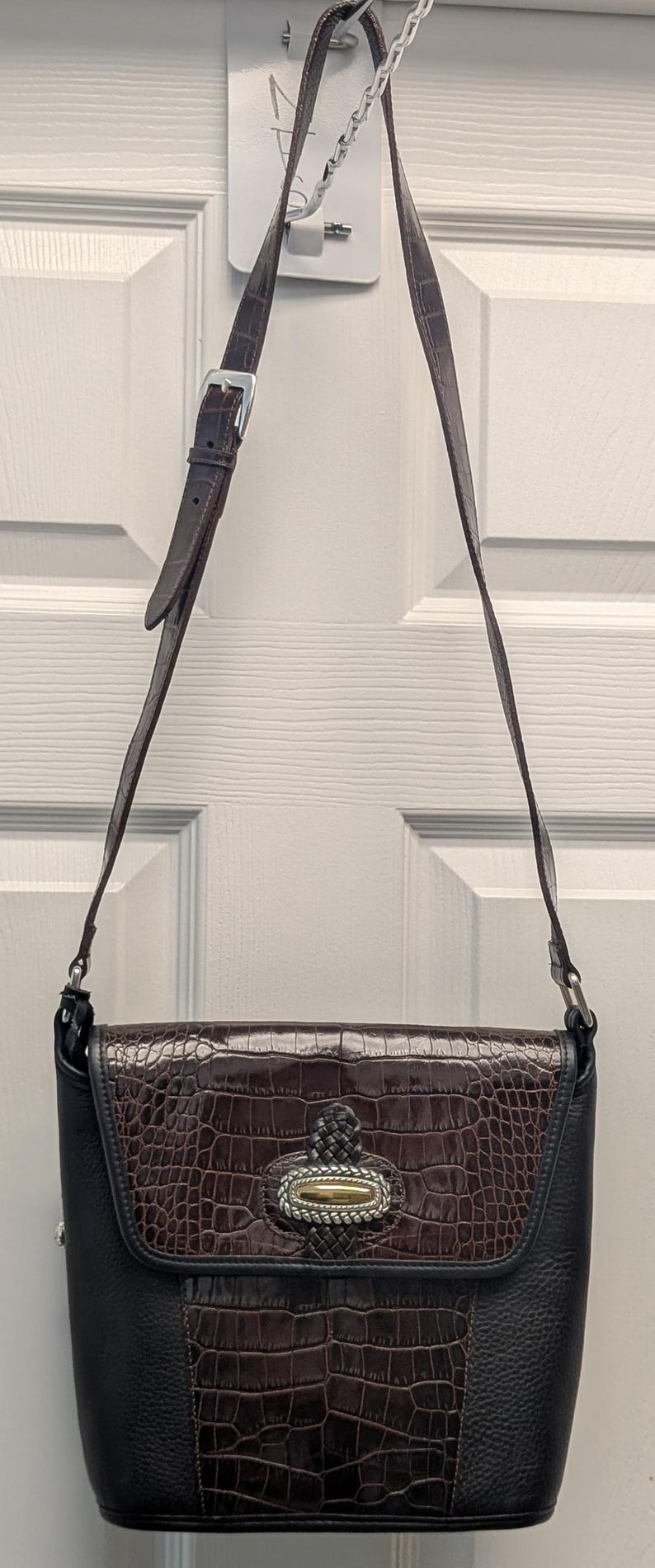 May include: A brown and black leather crossbody bag with a crocodile-textured flap. The bag has a silver buckle on the strap and a decorative silver and gold clasp. The bag is hanging from a white door.