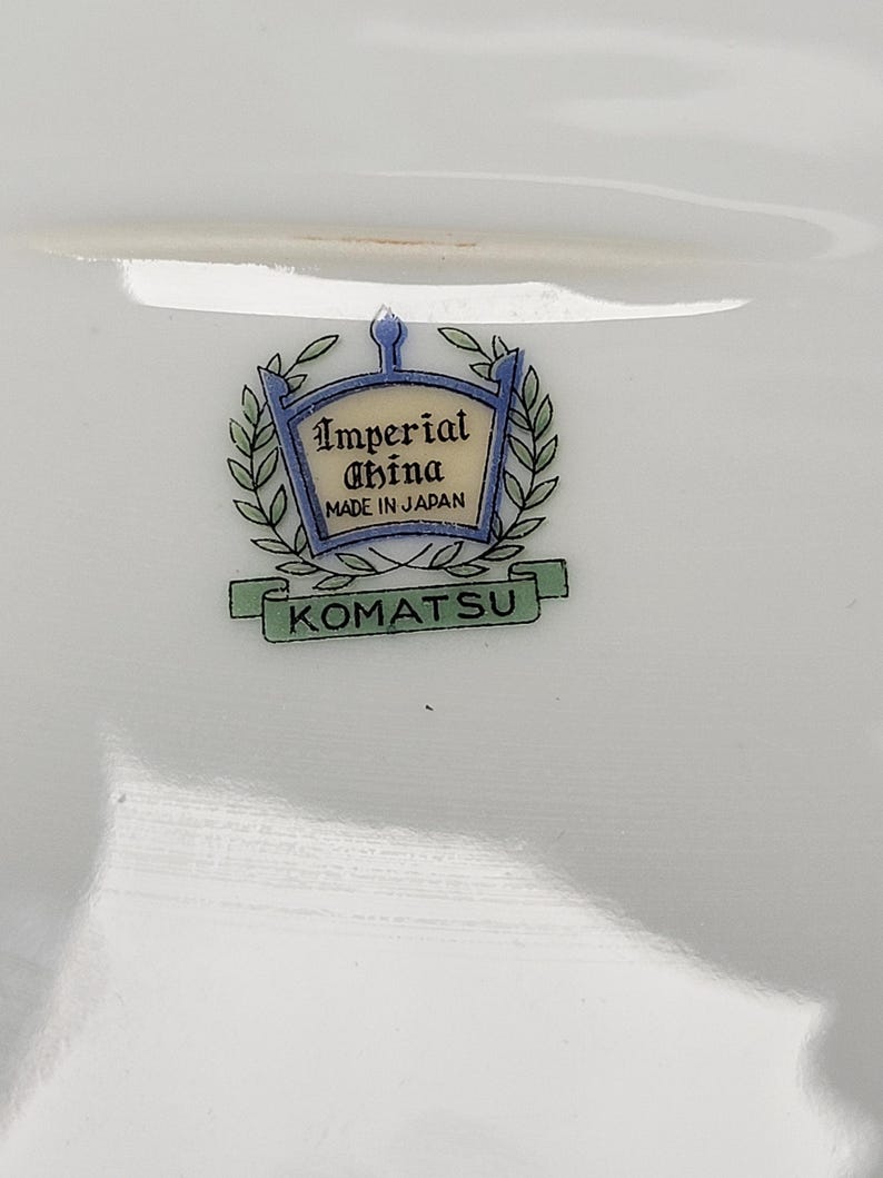 May include: White porcelain dish with the text "Imperial China MADE IN JAPAN" and the name "KOMATSU" in a green banner. The logo is framed by a blue and gold crest with laurel leaves.