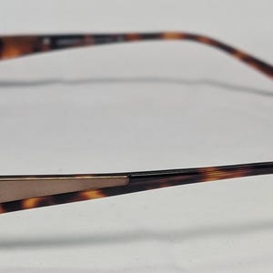 May include: Brown and gold-toned eyeglasses with a rectangular frame. The temples feature a tortoiseshell pattern and the brand name "Versace". The glasses have a classic, sophisticated design.