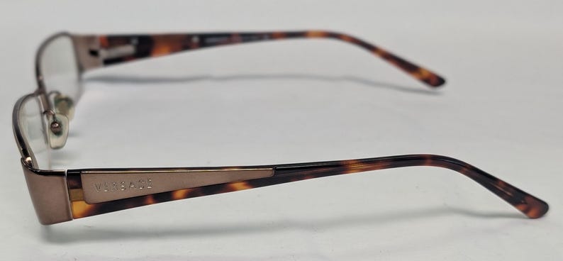 May include: Brown and gold-toned eyeglasses with a rectangular frame. The temples feature a tortoiseshell pattern and the brand name "Versace". The glasses have a classic, sophisticated design.