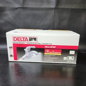 May include: A white box featuring a silver faucet on the front. The box is labelled "DELTA" and "Brilliance" with the model number "N544-NPWF". The faucet is a single-handle, chrome finish, and includes a pop-up drain. The box also includes a lifetime warranty.