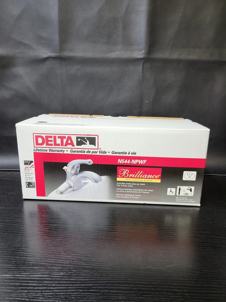 May include: A white box featuring a silver faucet on the front. The box is labelled "DELTA" and "Brilliance" with the model number "N544-NPWF". The faucet is a single-handle, chrome finish, and includes a pop-up drain. The box also includes a lifetime warranty.