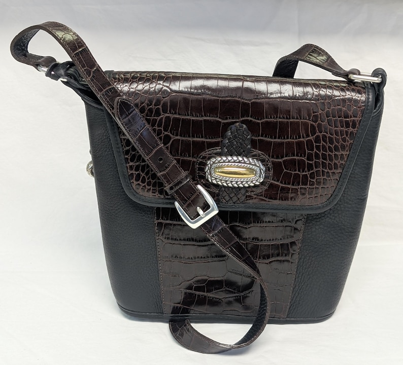 May include: A brown and black leather crossbody bag with a crocodile-textured flap and accents. The bag features a silver buckle and a decorative clasp with gold and silver details. The adjustable strap is also brown leather.