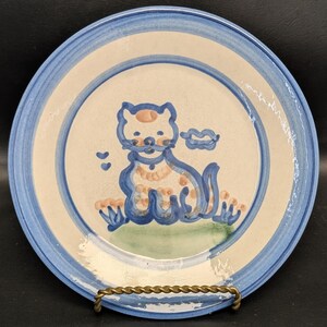 May include: A ceramic plate with a hand-painted cat design. The plate features a blue rim and a cream-colored center. The cat is depicted in blue and orange, with a green base and two small blue hearts. The plate is supported by a gold-colored metal stand.