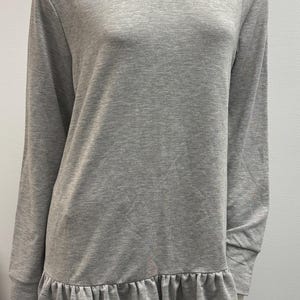 May include: A light gray long-sleeve top with a round neckline and a ruffled hem. The top is made of a soft, solid-colored fabric. The sleeves are long and end at the wrists.