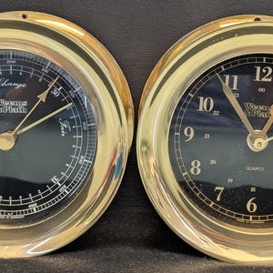 May include: Two round, gold-toned nautical instruments. One is a barometer with "Rain" and "Fair" indicators, the other a clock with a black face and gold hands. Both have the text "Weems & Plath".
