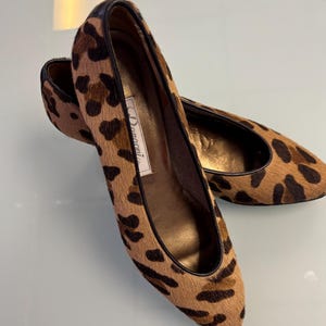 May include: A pair of brown and black leopard print flats with a pointed toe. The shoes are made of a soft, hairy material and have a black leather trim. The brand name "Damiani" is printed on the inside of the shoe.