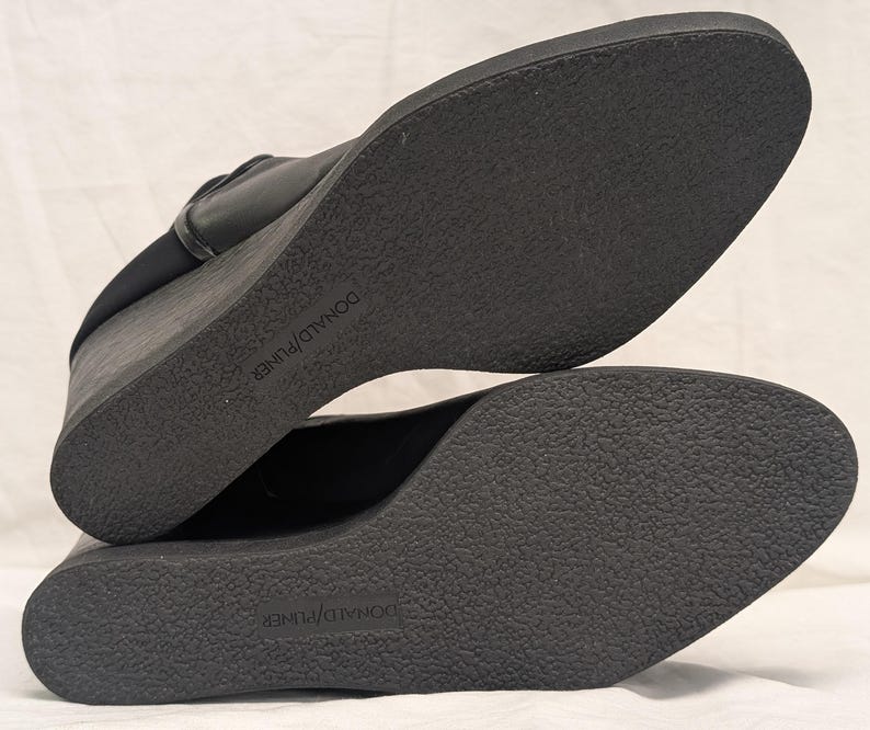 May include: Black wedge-heeled shoes with textured soles. The soles are marked with the brand name "DONALD J PLINER". The shoes have a sleek, modern design, suitable for various occasions.