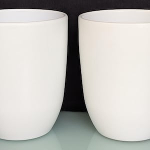 May include: Two white ceramic cups with a smooth, matte finish. The cups have a rounded base and a slightly flared rim. They are set against a black background.