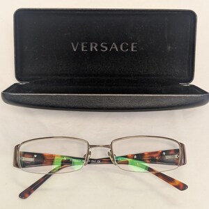 May include: Versace eyeglasses with a brown and gold frame and tortoise shell accents. The glasses are displayed with an open black case with the Versace logo.