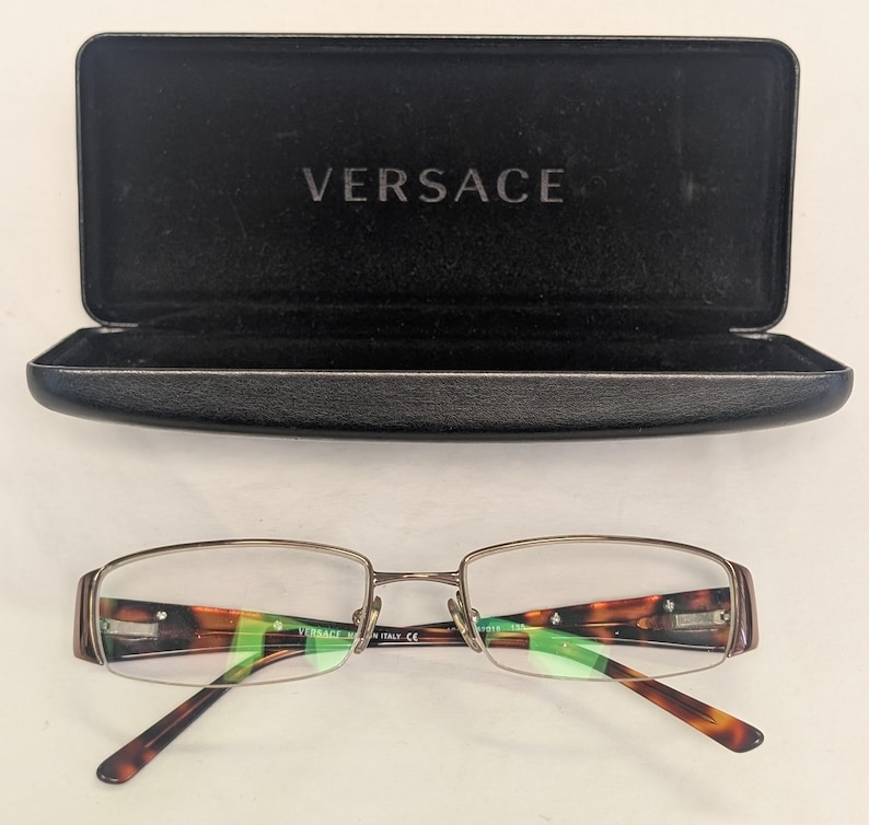 May include: Versace eyeglasses with a brown and gold frame and tortoise shell accents. The glasses are displayed with an open black case with the Versace logo.