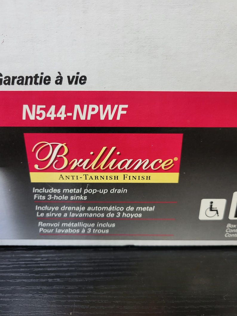 May include: A red and black box with the text "Garantie &agrave; vie" and "N544-NPWF" on the top. The box also features the text "Brilliance Anti-Tarnish Finish" and includes details about the product's features, such as a metal pop-up drain and a 3-hole sink.