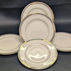 May include: A collection of six white ceramic plates with a gold and gray border. The plates are arranged in a circular pattern, showcasing their elegant design. The plates have a simple, yet sophisticated aesthetic, suitable for formal dining.