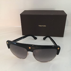 May include: Black sunglasses with gold accents and a Tom Ford logo on the case. The sunglasses have a dark gray lens and a thick black frame.