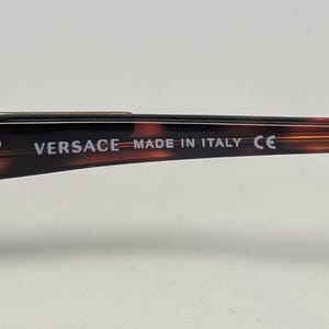 May include: Close-up of the arm of a pair of eyeglasses with a tortoiseshell pattern. The text "VERSACE MADE IN ITALY CE" is printed in white on the arm of the glasses. A small silver stud is visible near the text.