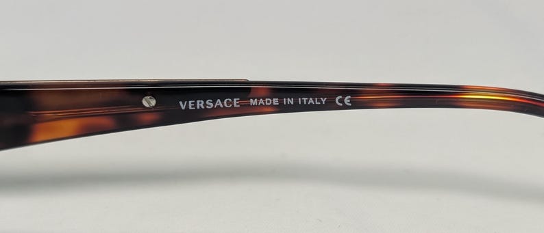 May include: Close-up of the arm of a pair of eyeglasses with a tortoiseshell pattern. The text "VERSACE MADE IN ITALY CE" is printed in white on the arm of the glasses. A small silver stud is visible near the text.