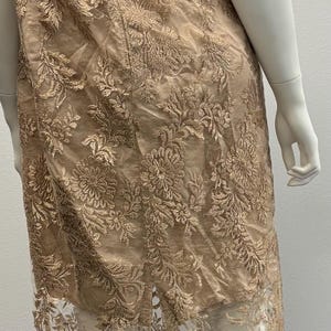 Pode incluir: Um vestido comprido, sem ombros, com um padr&atilde;o de renda floral em cor castanho claro. O vestido tem uma sobreposi&ccedil;&atilde;o transparente e um corpete justo.