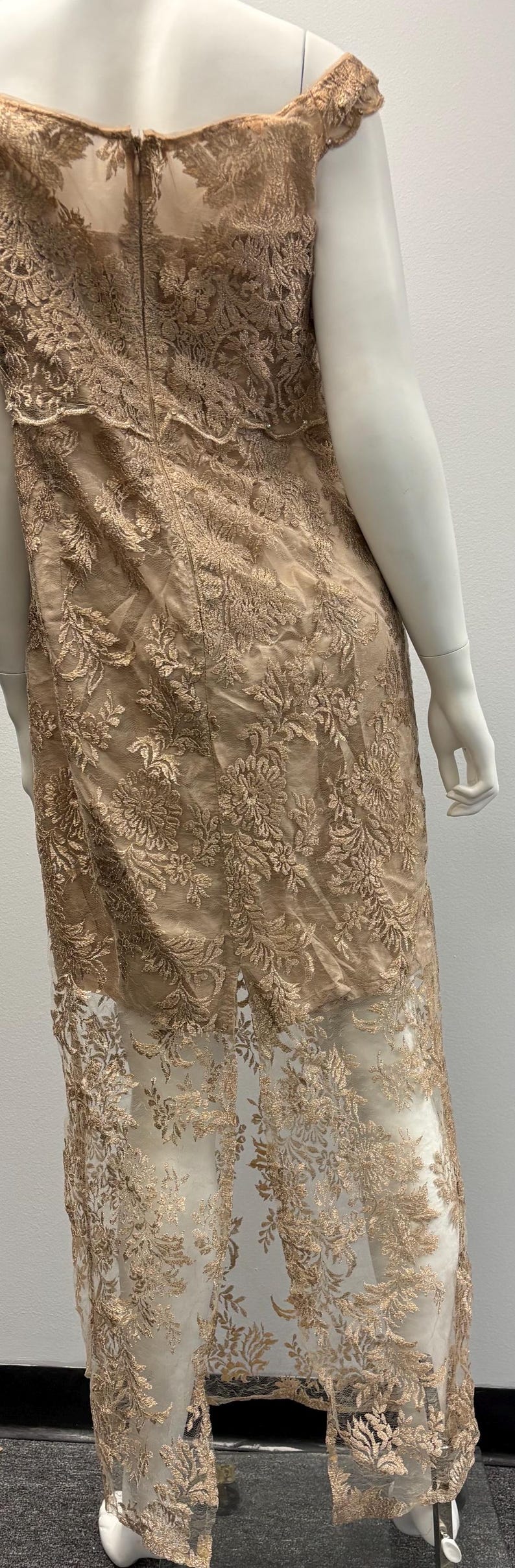 Pode incluir: Um vestido comprido, sem ombros, com um padr&atilde;o de renda floral em cor castanho claro. O vestido tem uma sobreposi&ccedil;&atilde;o transparente e um corpete justo.