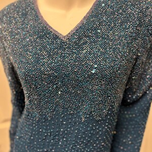 May include: Teal-colored beaded top with a V-neckline. The top features long sleeves and is heavily embellished with small beads and sequins, creating a textured, shimmering effect. The neckline is trimmed with a contrasting lavender-colored beading.