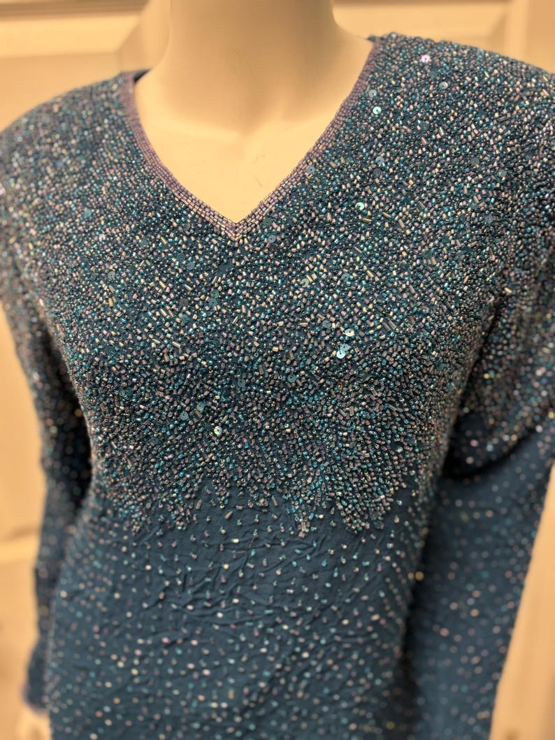 May include: Teal-colored beaded top with a V-neckline. The top features long sleeves and is heavily embellished with small beads and sequins, creating a textured, shimmering effect. The neckline is trimmed with a contrasting lavender-colored beading.