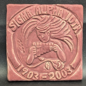May include: A square, reddish-brown tile with raised relief details. The tile features the words "SIGMA ALPHA IOTA" in an arch, a figure playing a pan flute, and the dates "1903=2003".