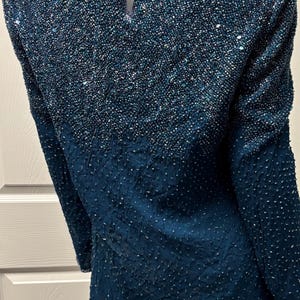 May include: A teal-colored, long-sleeved top with a beaded design. The upper portion of the top is densely covered in small, iridescent beads, creating a shimmering effect. The lower section has a textured pattern. The neckline is trimmed with a delicate beaded border.