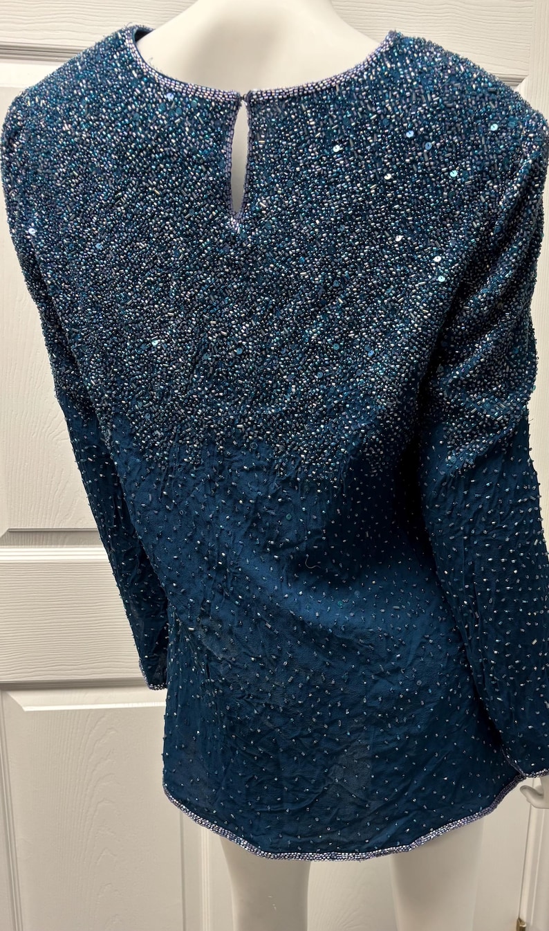 May include: A teal-colored, long-sleeved top with a beaded design. The upper portion of the top is densely covered in small, iridescent beads, creating a shimmering effect. The lower section has a textured pattern. The neckline is trimmed with a delicate beaded border.