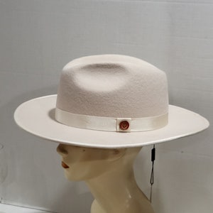 May include: A cream-colored fedora hat with a wide brim and a white ribbon band. The hat has a small, round, brown logo on the ribbon.