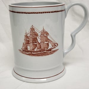 May include: White ceramic mug with a brown illustration of a sailing ship. The mug has a curved handle and a brown rope design around the rim. The base and rim of the mug are also trimmed in brown.