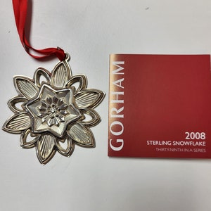 May include: A silver snowflake ornament with a red ribbon. The ornament is part of a series and is labeled "2008 Sterling Snowflake Thirty-ninth in a Series" and "GORHAM".