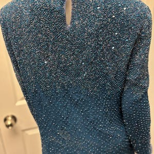 May include: A teal-colored beaded top with a keyhole back. The top is covered in small beads that create a textured surface. The sleeves are long and the neckline is round. The beads transition from a lighter shade at the top to a darker shade at the bottom.