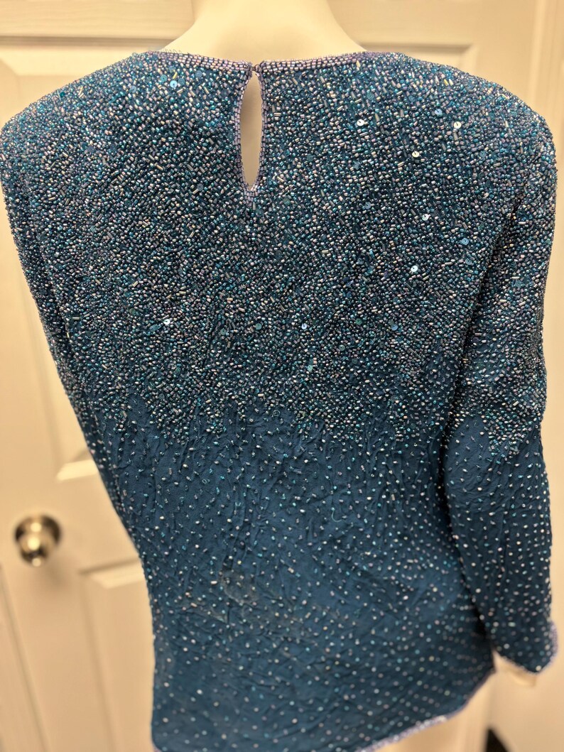 May include: A teal-colored beaded top with a keyhole back. The top is covered in small beads that create a textured surface. The sleeves are long and the neckline is round. The beads transition from a lighter shade at the top to a darker shade at the bottom.