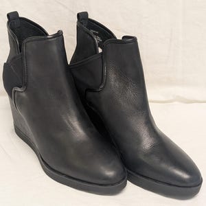 May include: A pair of black leather wedge ankle boots. The boots have a sleek design with a rounded toe, elastic side panels, and a wedge heel. The boots are made of smooth leather and have a rubber sole.