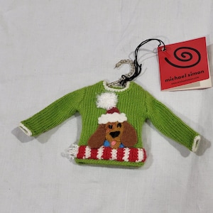 May include: A green knitted sweater ornament with a dog wearing a Santa hat. The dog has brown fur and a blue tongue. The sweater has a white pom-pom and a red and white striped scarf. A red tag with the words "michael simon" is attached.