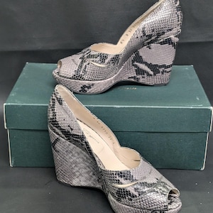 May include: A pair of gray and black snakeskin-patterned wedge heel shoes with open toes. The shoes are on top of a green box. The heels have a platform sole and a peep-toe design. The shoes are a size 8.5.