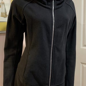 May include: Black zip-up jacket with a mock neck and a long, fitted style. The jacket has a silver zipper and a quilted detail on the bottom.
