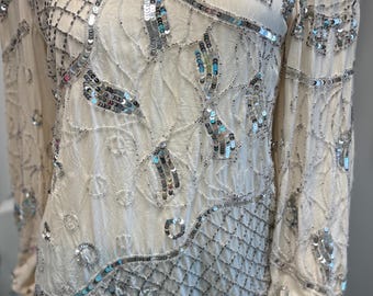 May include: Cream-colored long-sleeved top with intricate silver sequin and bead embellishments. The design features geometric patterns and floral motifs, creating a vintage, elegant look. The top is paired with a green skirt, suggesting a complete outfit.