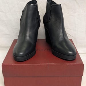 May include: A pair of black leather ankle boots with wedge heels. The boots are displayed on a red box. The boots have a sleek design with a rounded toe and elastic side panels. The box has the text "ALD / PLI" printed on it.