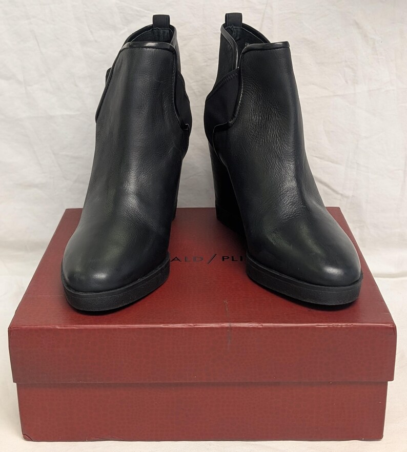 May include: A pair of black leather ankle boots with wedge heels. The boots are displayed on a red box. The boots have a sleek design with a rounded toe and elastic side panels. The box has the text "ALD / PLI" printed on it.