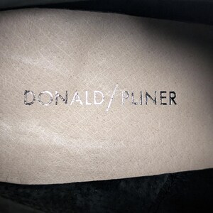 May include: Close-up of the interior of a shoe with the brand name "DONALD/PLINER" in silver lettering on a beige leather insole. The shoe's interior is visible, with a black suede exterior.